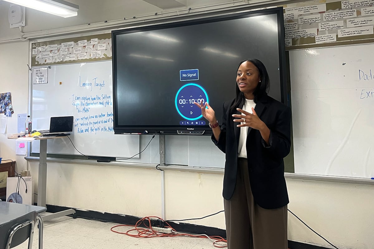 A woman in a dark suit jacket speaks in front of whiteboards.