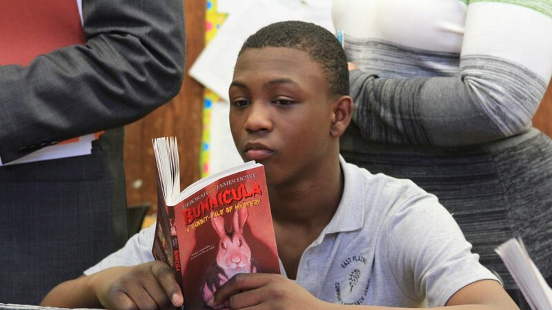 A boy in a blue school uniform sits at a table reading a book.