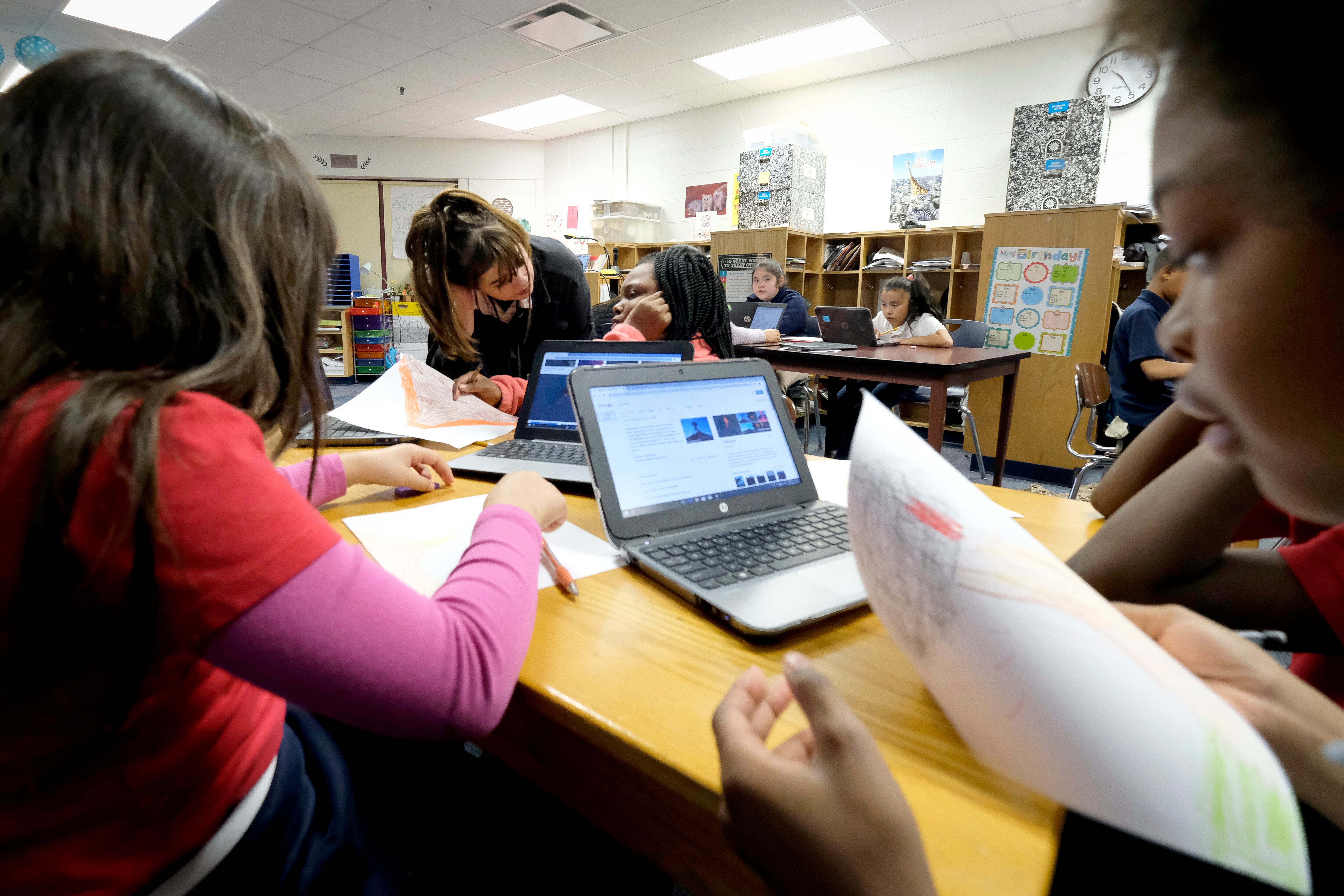 Students sitting around a table look at worksheets, with a laptop open in front of them. A student rests her face in her hand while a teacher leans down to talk to her.