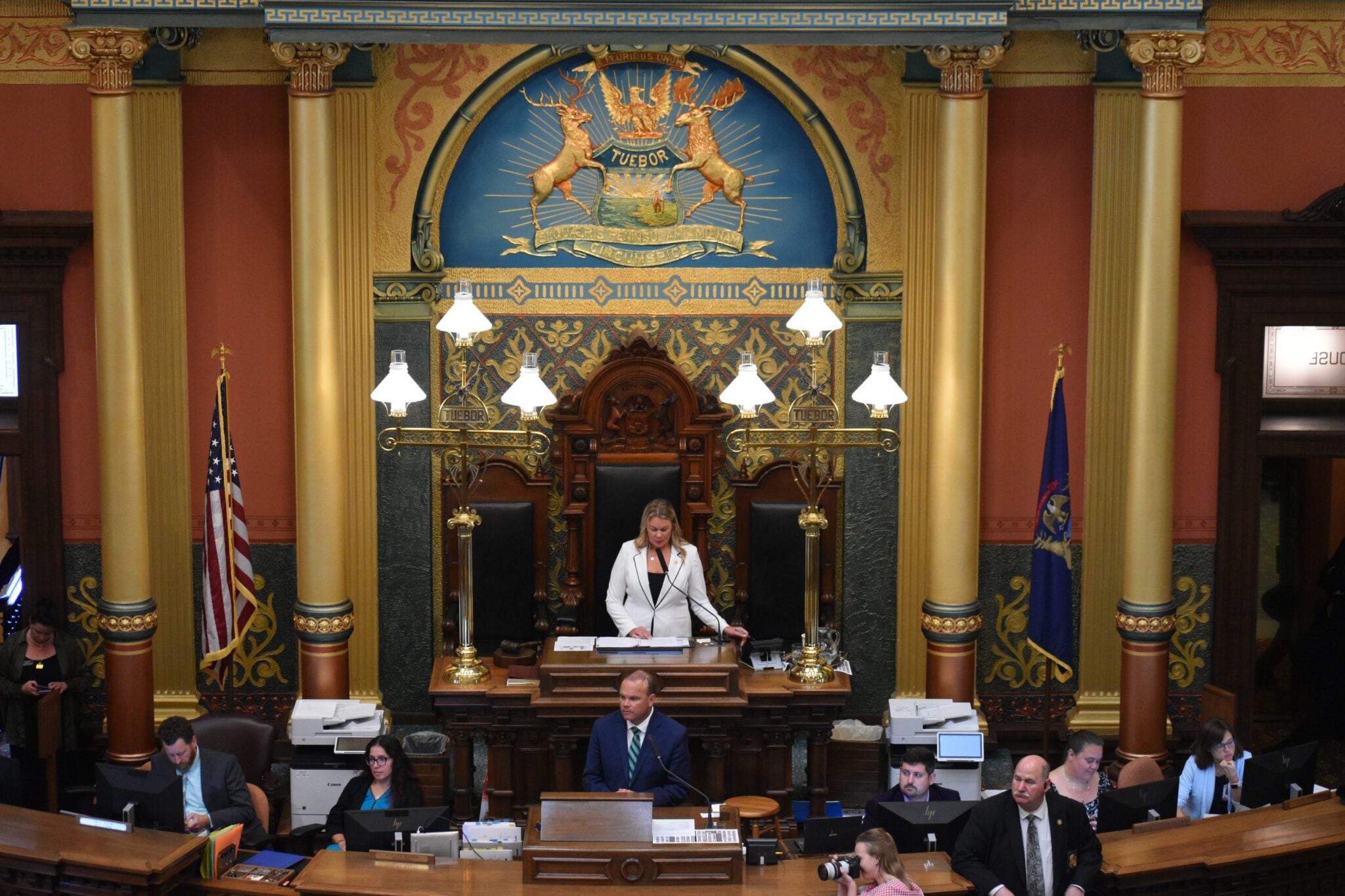 A photograph of a large group of people in a large state capitol room with flags and ornate decorations.