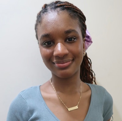 A high school student with long dark braids and wearing a gold name plate necklace poses for a portrait in front of a white wall.