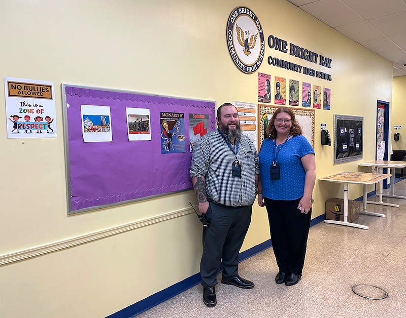 A man and woman stand in front of a purple bulletin board.