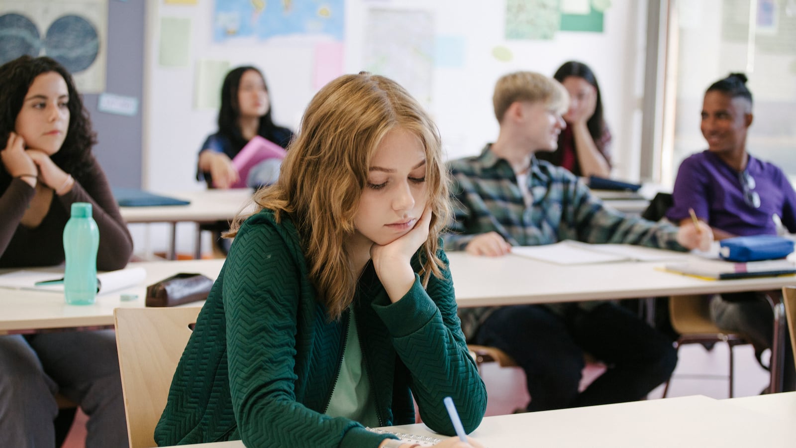 A female student wearing a green sweater takes notes while working in a classroom with her classmates.