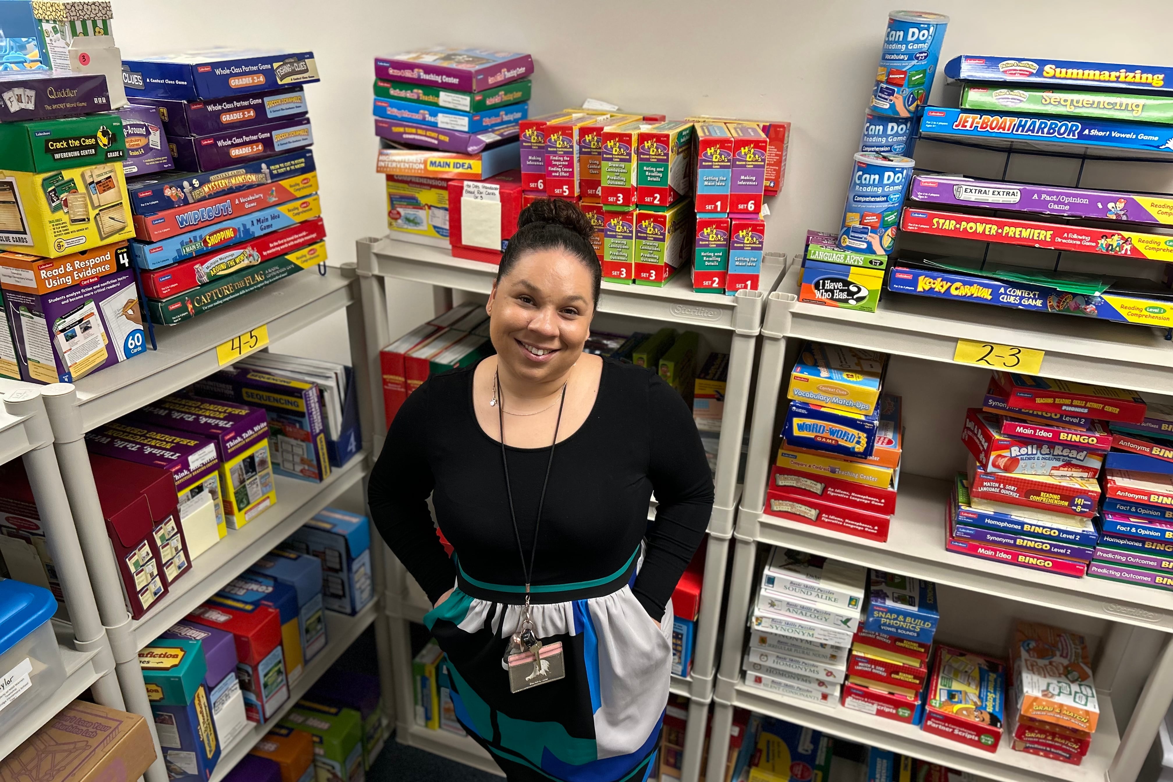 A woman in a black, green, and blue dress smiles while standing in front of shelves with children’s games on them.