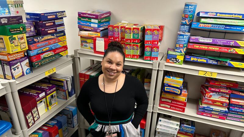 A woman in a black, green, and blue dress smiles while standing in front of shelves with children’s games on them.
