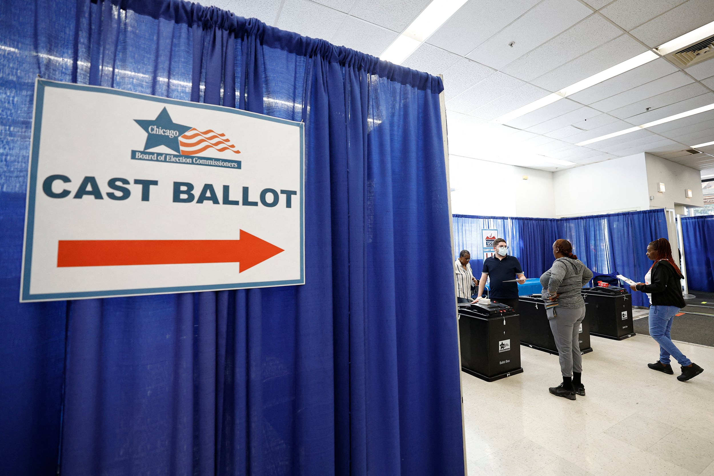 A large sign that reads "Cast Ballot" hangs on a blue curtain in a room with a couple of people in the background working voting machines.