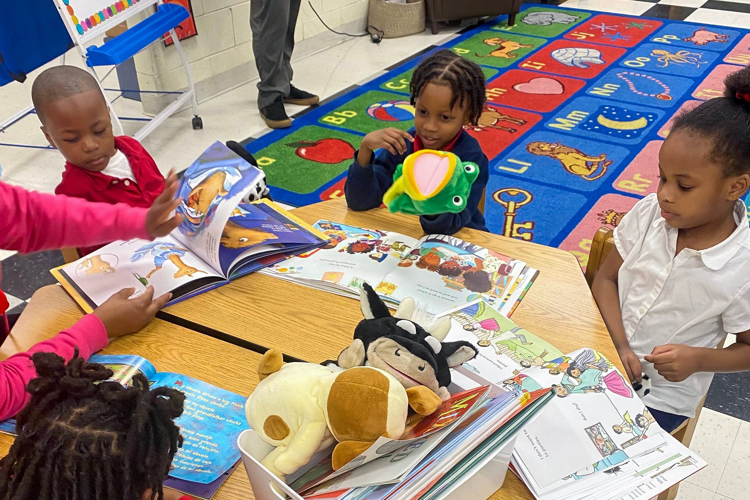 Three kindergarten read books together with their teacher at a table with books and hand puppets of different animals.