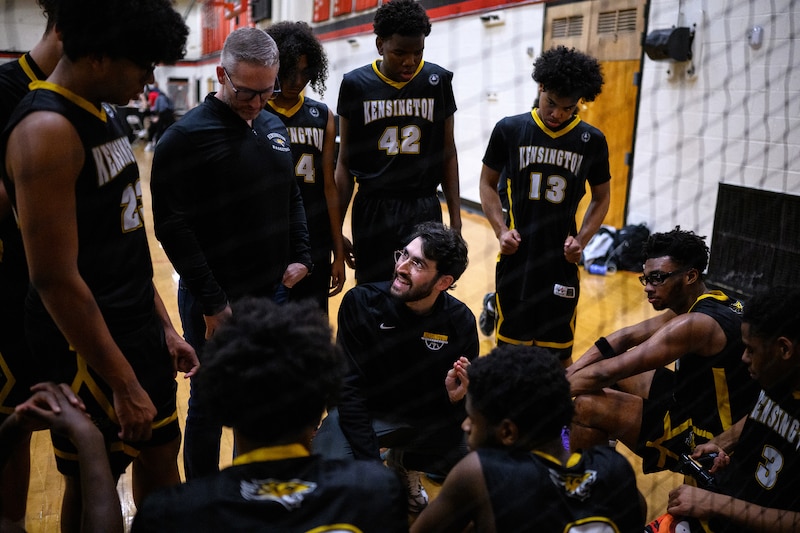 A photograph of a high school basketball team during a timeout in a school gym.