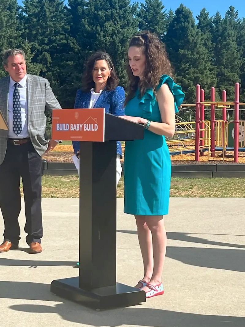 A woman in a blue dress speaks from a podium outside on a sunny day with two people in the background.