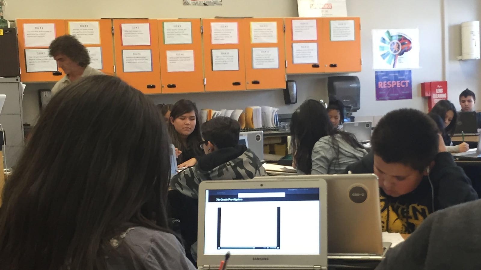 Students sit at wooden desks in a classroom with orange cabinets in the background.