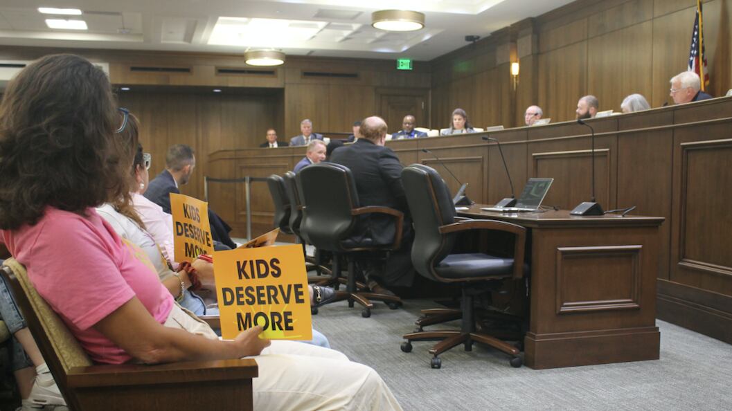 Several people hold signs that say “kids deserve more” in a legislative hearing room filled with people.
