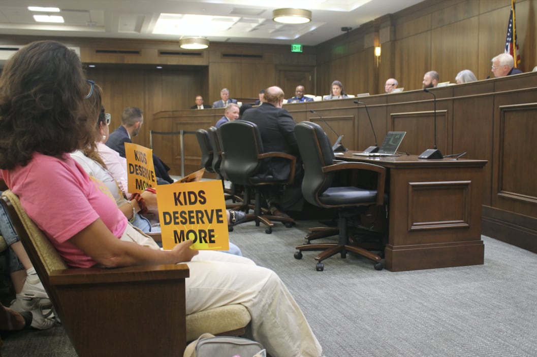 Several people hold signs that say “kids deserve more” in a legislative hearing room filled with people.