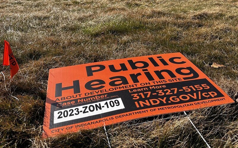 A red and black sign lays on grass with a red flag to the left of it. Large black words read "Public Hearing" with lots of smaller words.