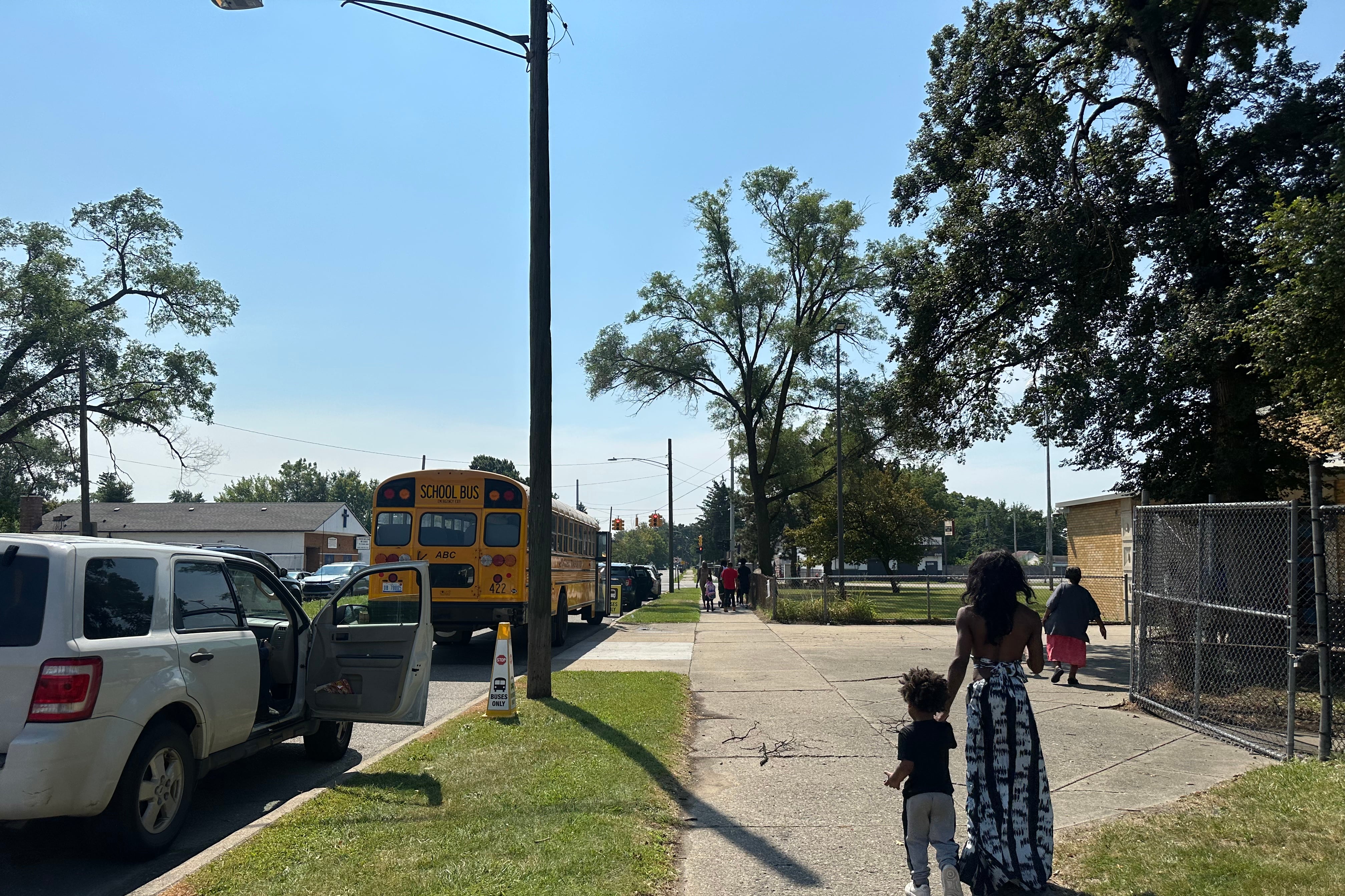 A woman walks down the street with her 3-year-old child. A yellow school bus is parking on the street.
