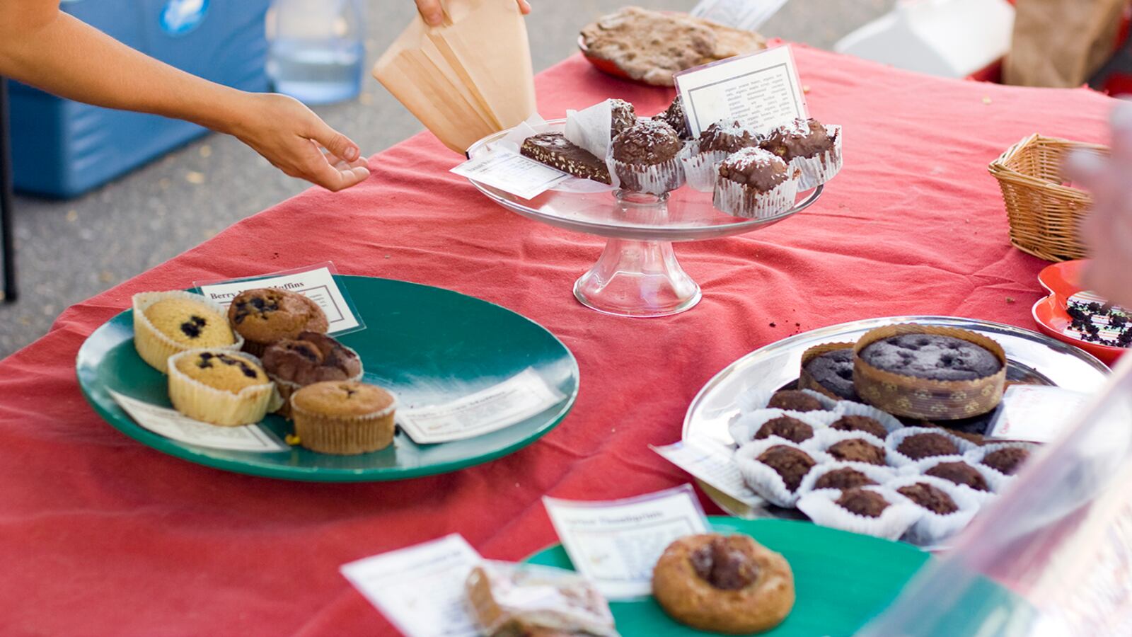 One hand holds open a pastry bag while the other hand reaches for a sweet treat on a table full of baked goods with a red table cloth.