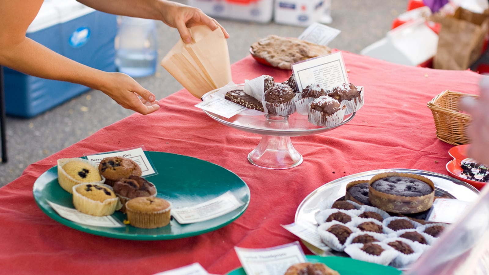 One hand holds open a pastry bag while the other hand reaches for a sweet treat on a table full of baked goods with a red table cloth.