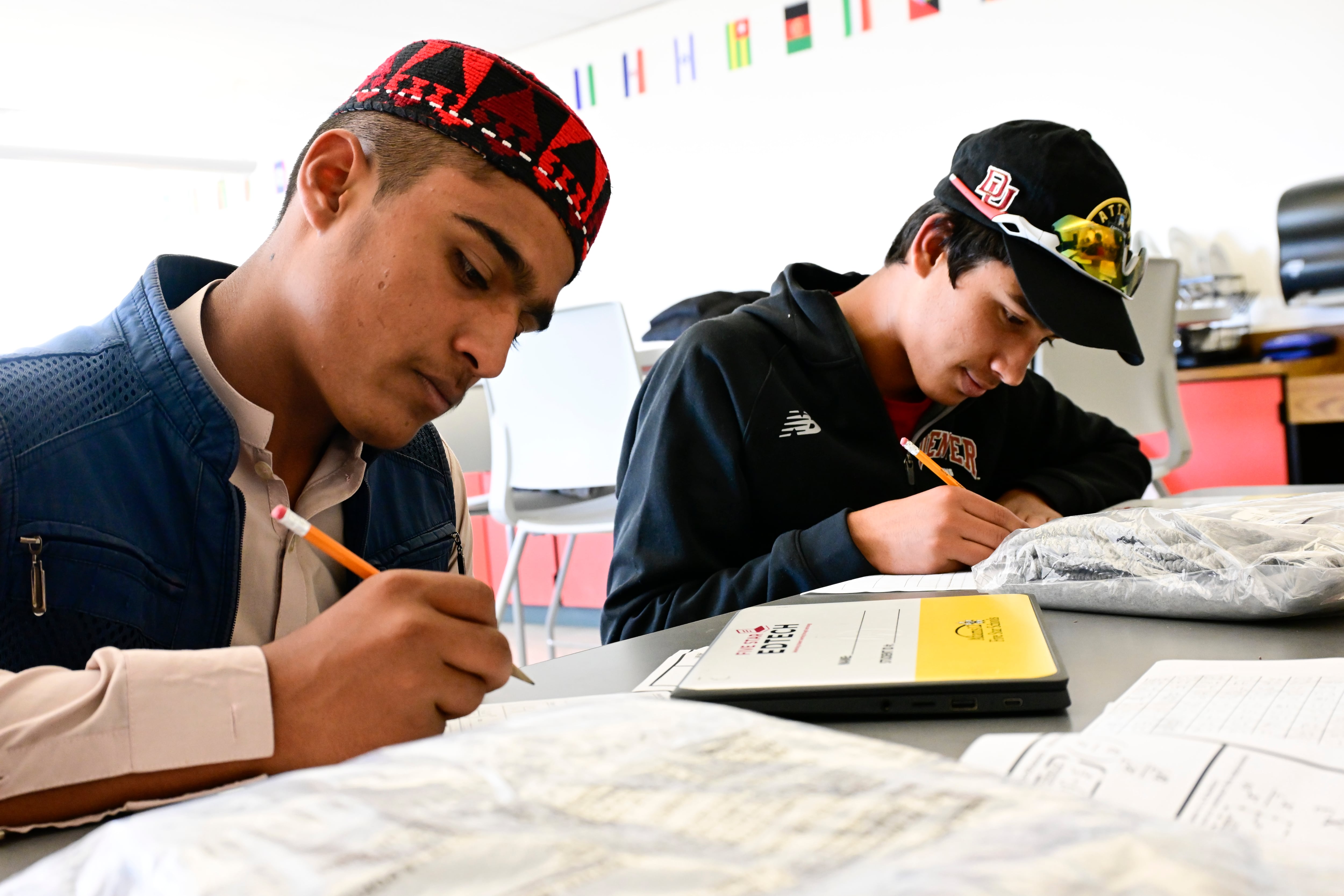 Two adolescent boys seated a school table holding pencils and writing on paper.