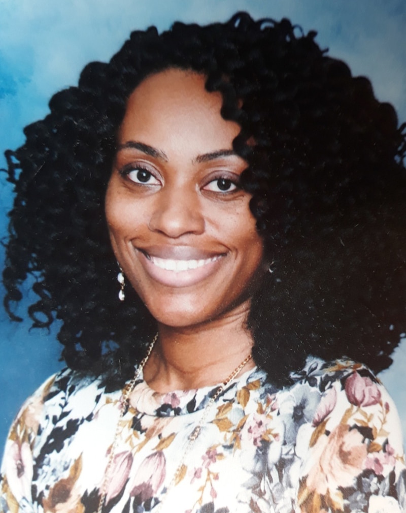 A portrait of a woman with short, dark, curly hair and wearing a floral blouse with a blue background.