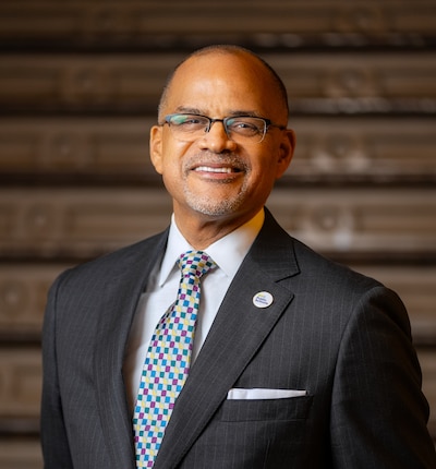 A man wearing a dark suit and tie and wearing classes smiles for a portrait in front of a dark background.