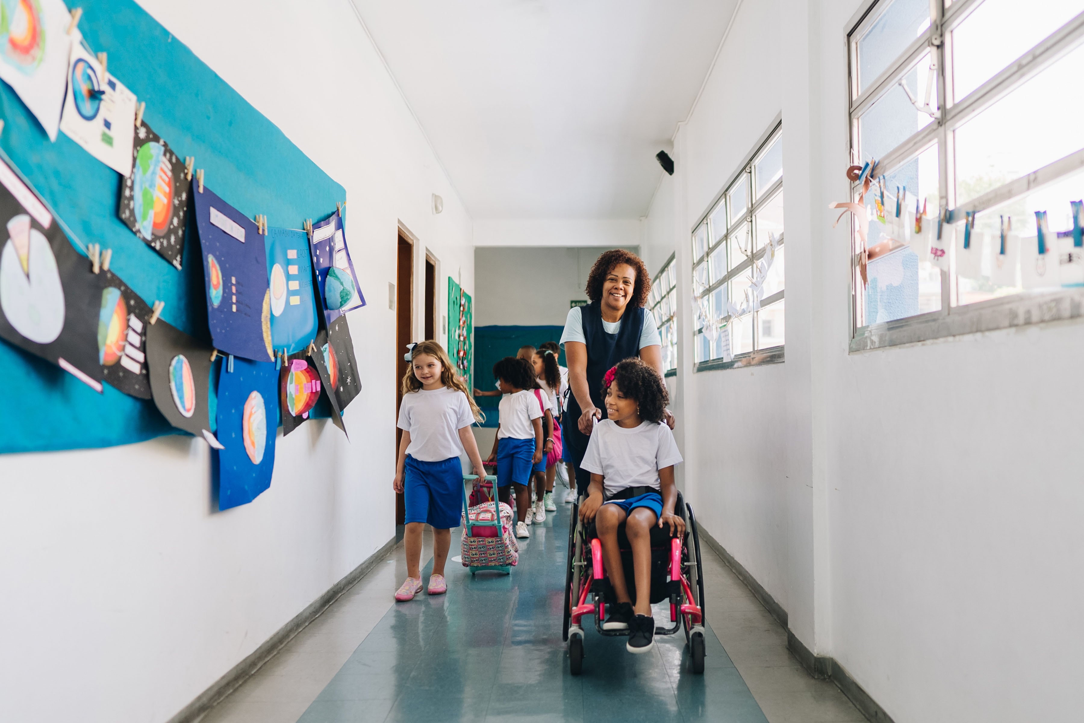 Teacher pushing student using wheelchair down a hallway at a school with other students in the background.