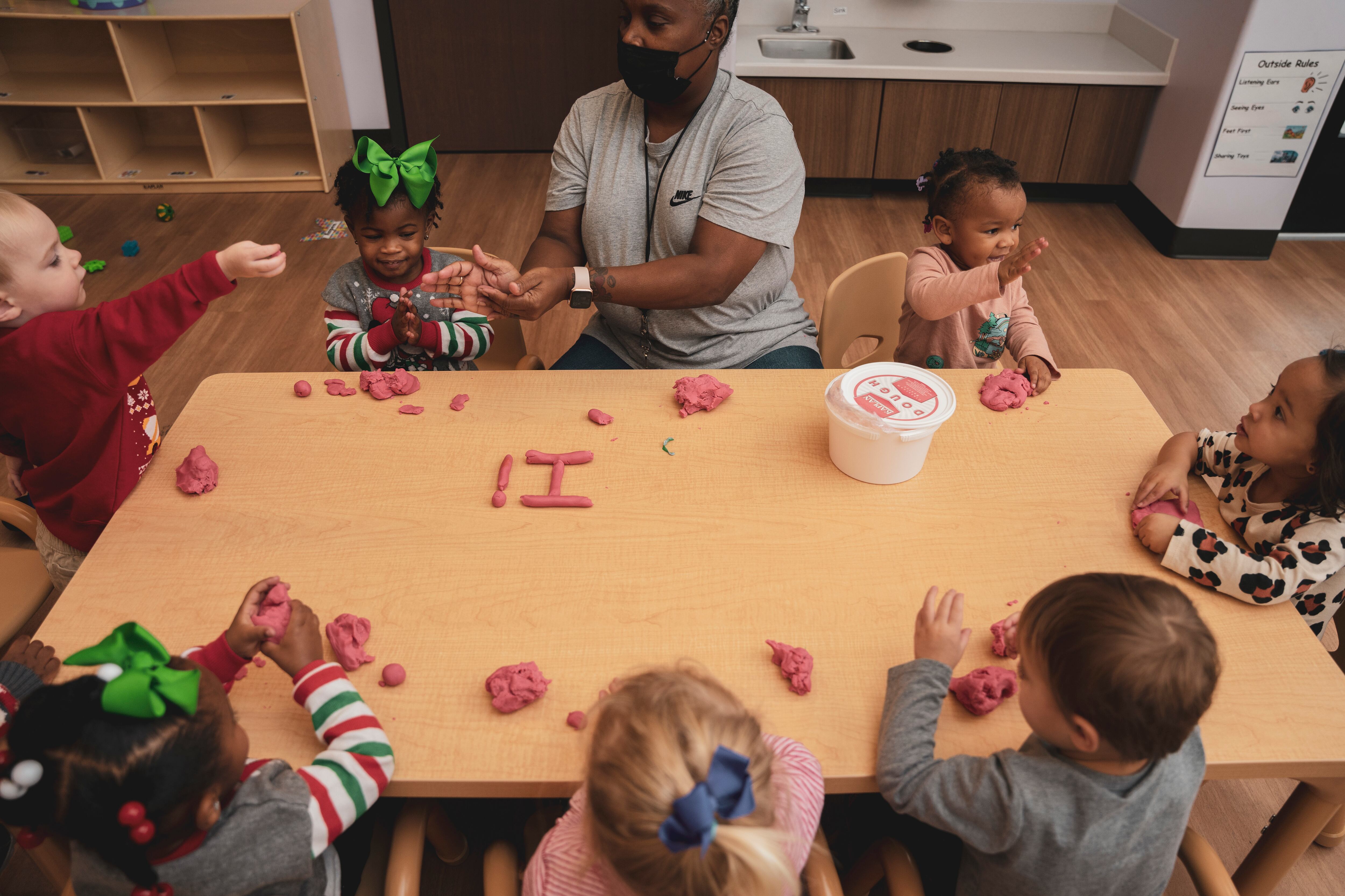 A preschool teacher at a table surrounded by children uses Play-Doh to shape letters of the alphabet.