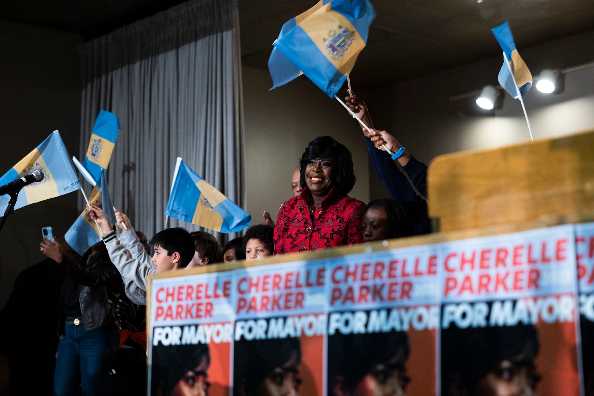 A woman wearing a red suit stands between a group of people waving blue and white flags on stage.