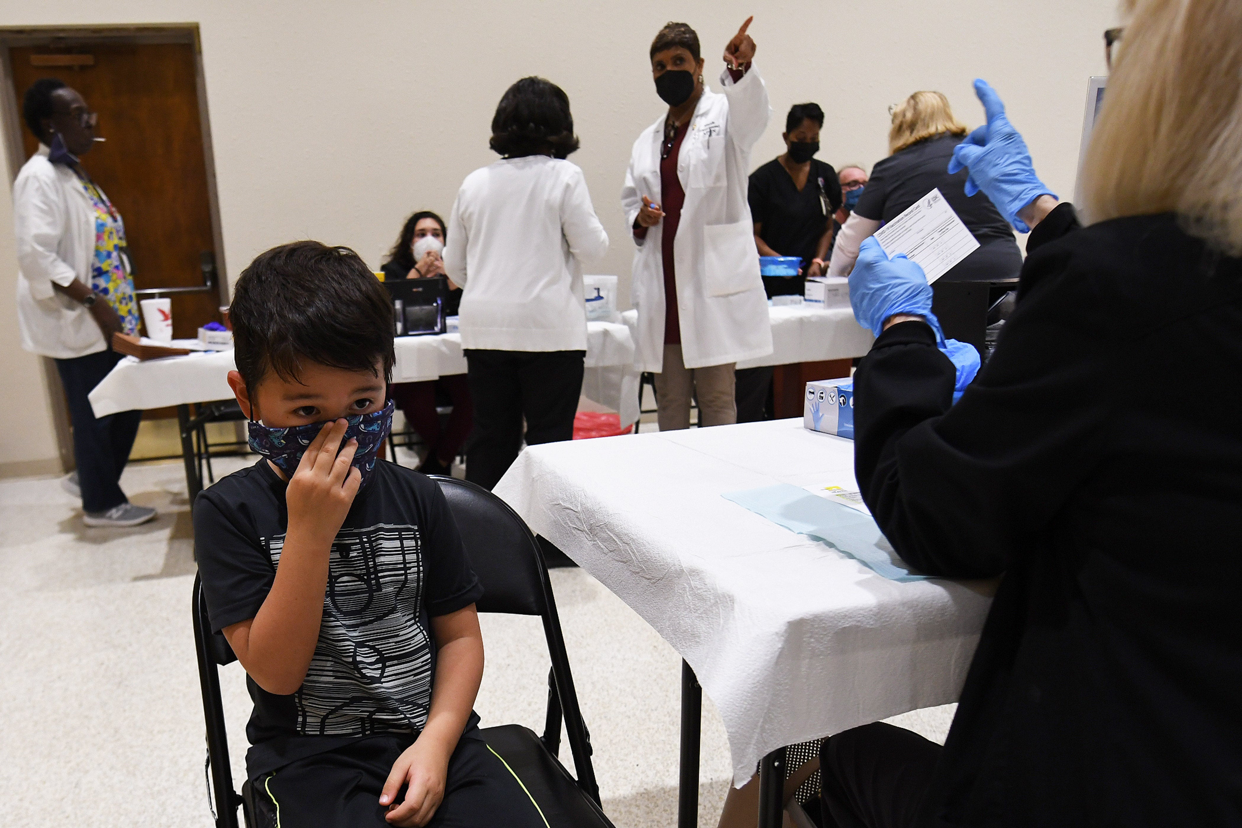A young boy wearing a black t-shirt prepares for a health care professional to administer a dose of a COVID vaccine, as others work at tables in the background.