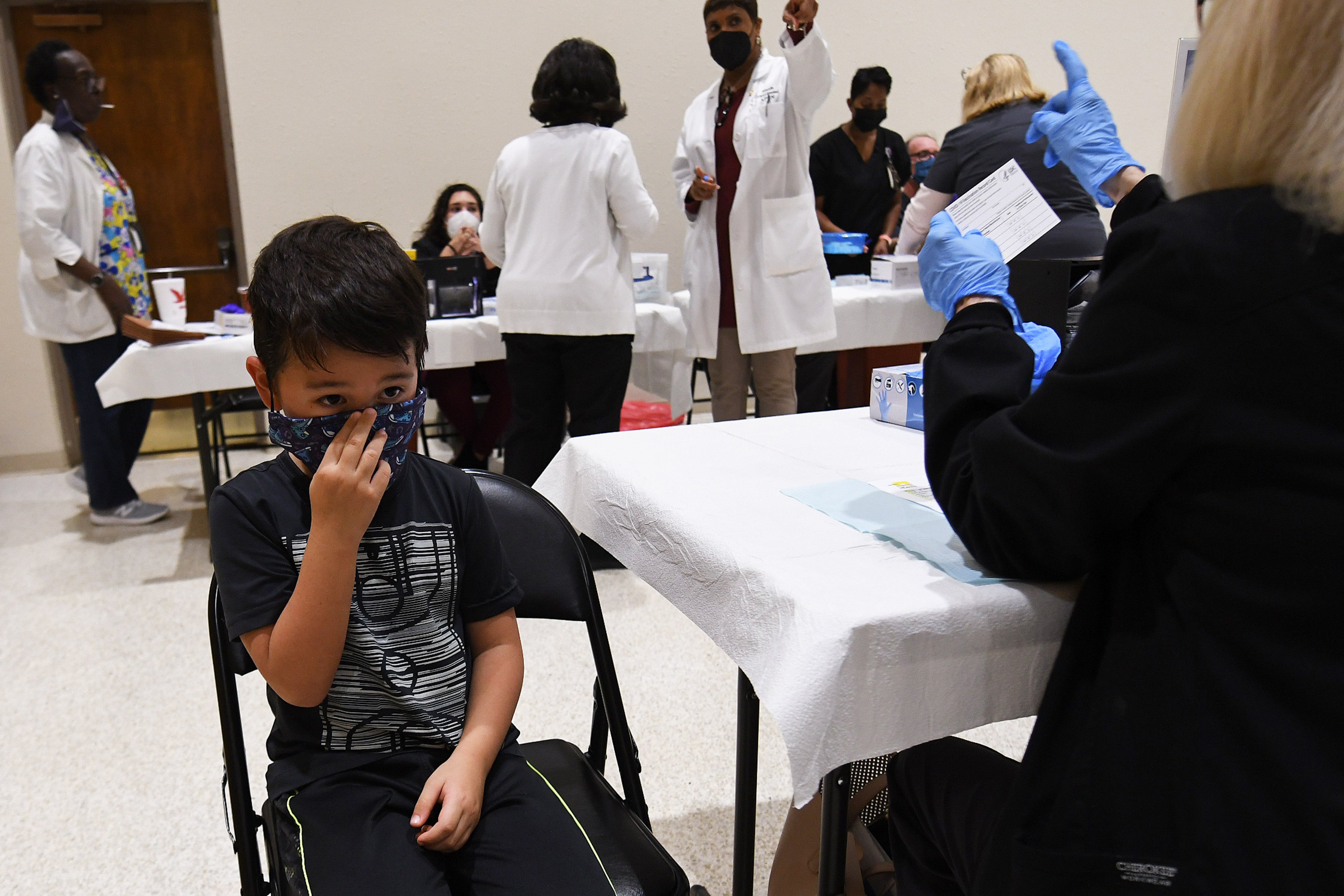 A young boy wearing a black t-shirt prepares for a health care professional to administer a dose of a COVID vaccine, as others work at tables in the background.