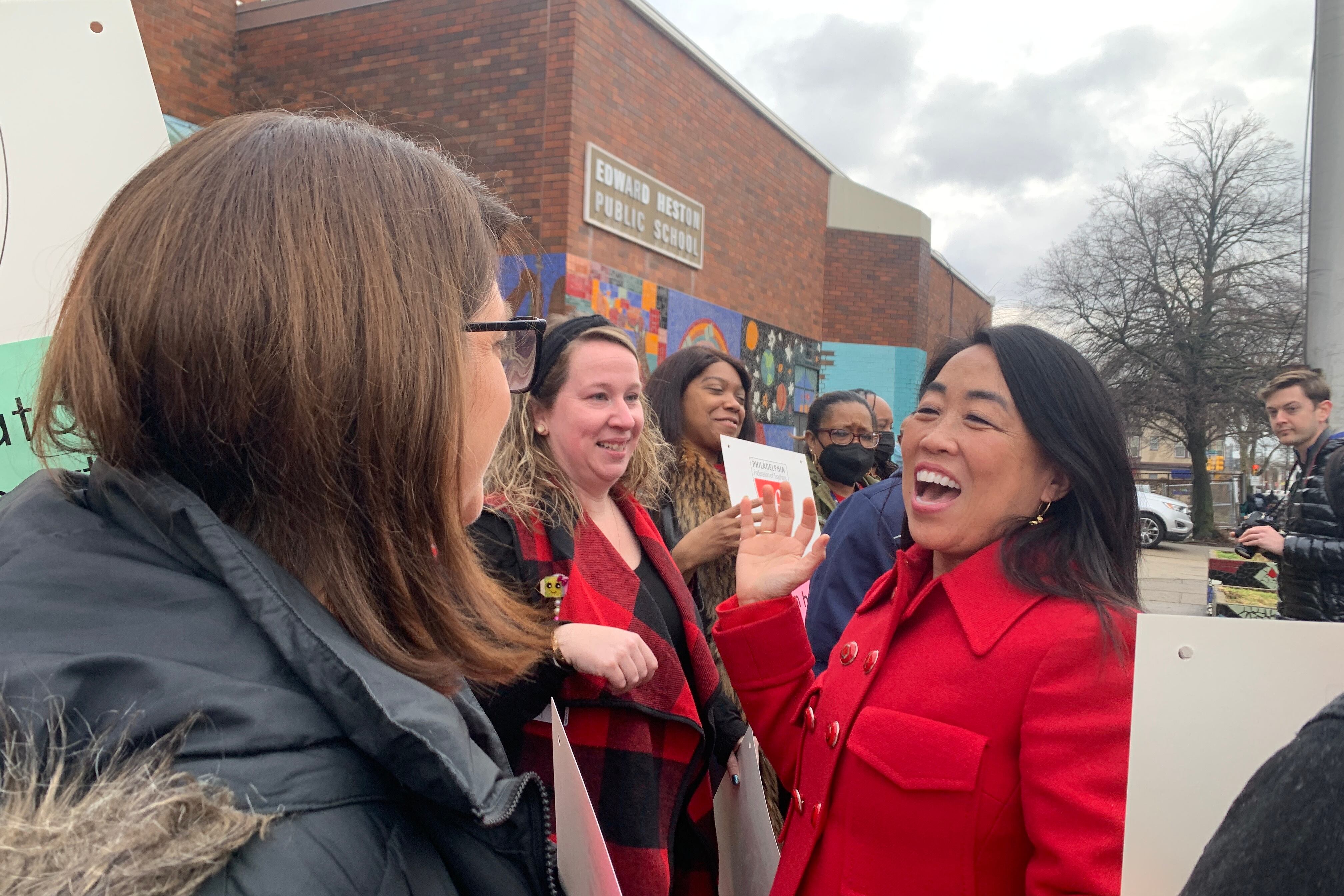 A woman with black hair wearing a red coat raises her right hand and laughs with other people in a crowd outside. In the background is a brick building with a sign reading “Edward Heston Public School.”