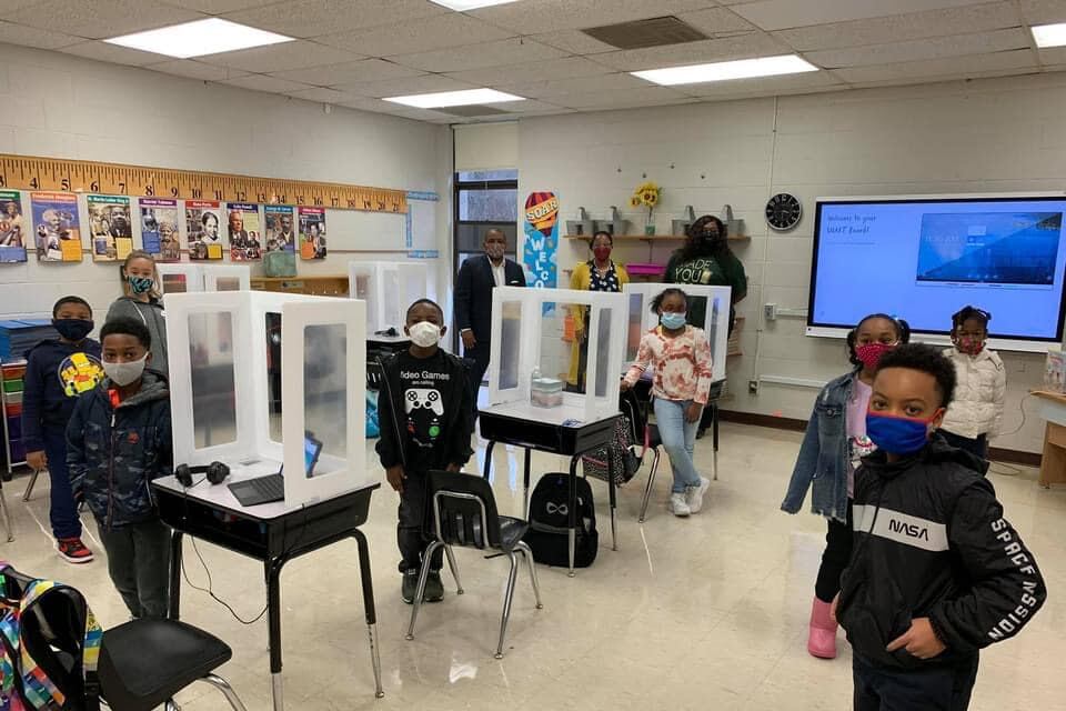 Students in masks pose standing next to their desks in a classroom inside of a school