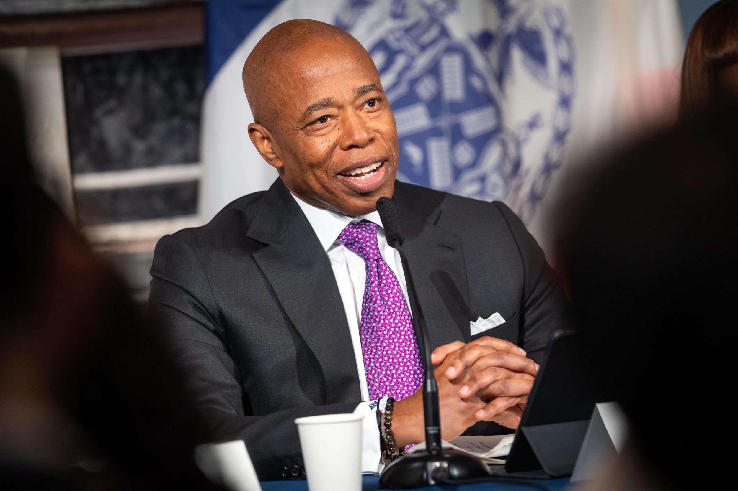 A man in a dark suit with a purple tie sits with folded hands