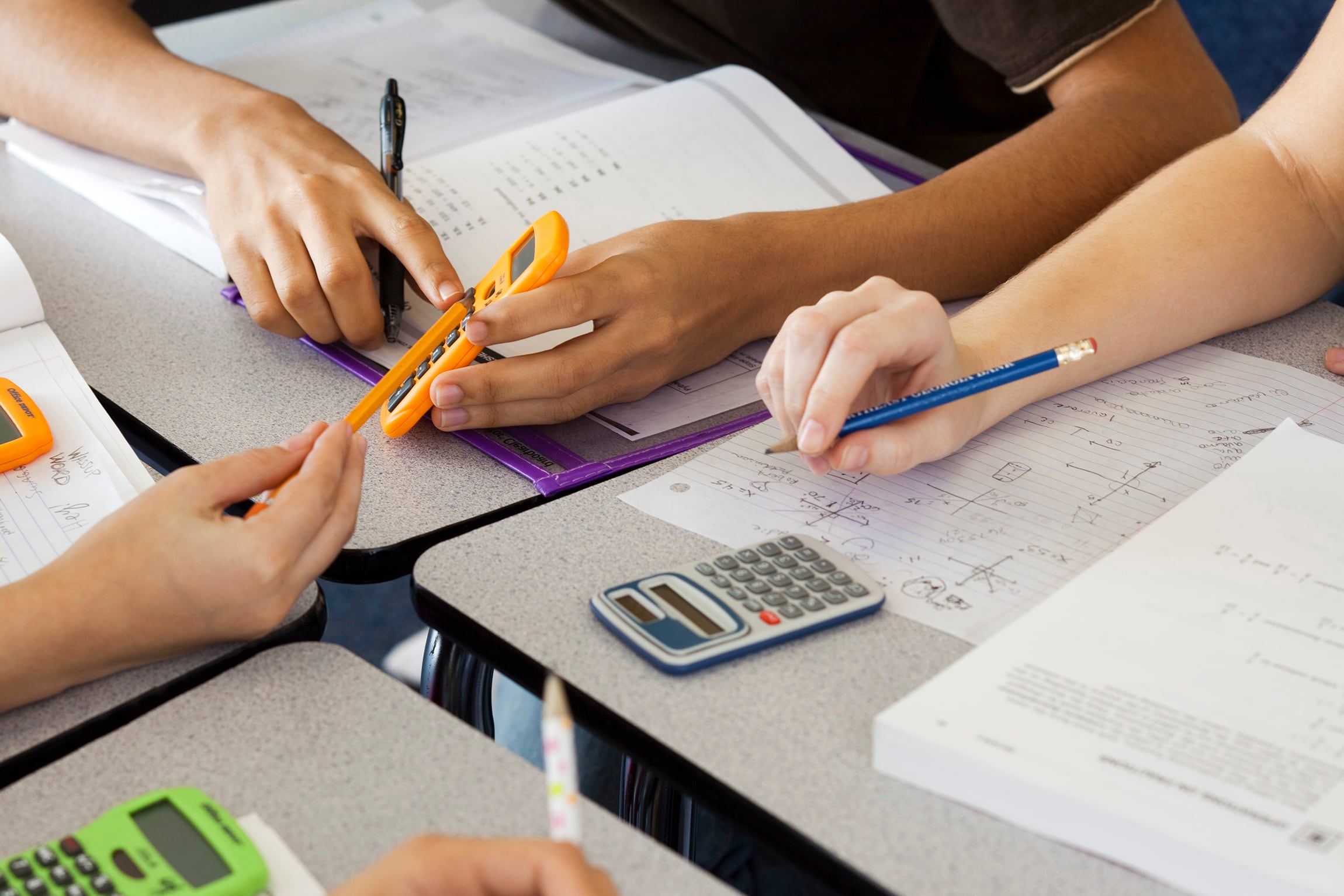 An up close photograph of three students working on math at their desks showing only their hands, white notebooks, pencils and calculators.