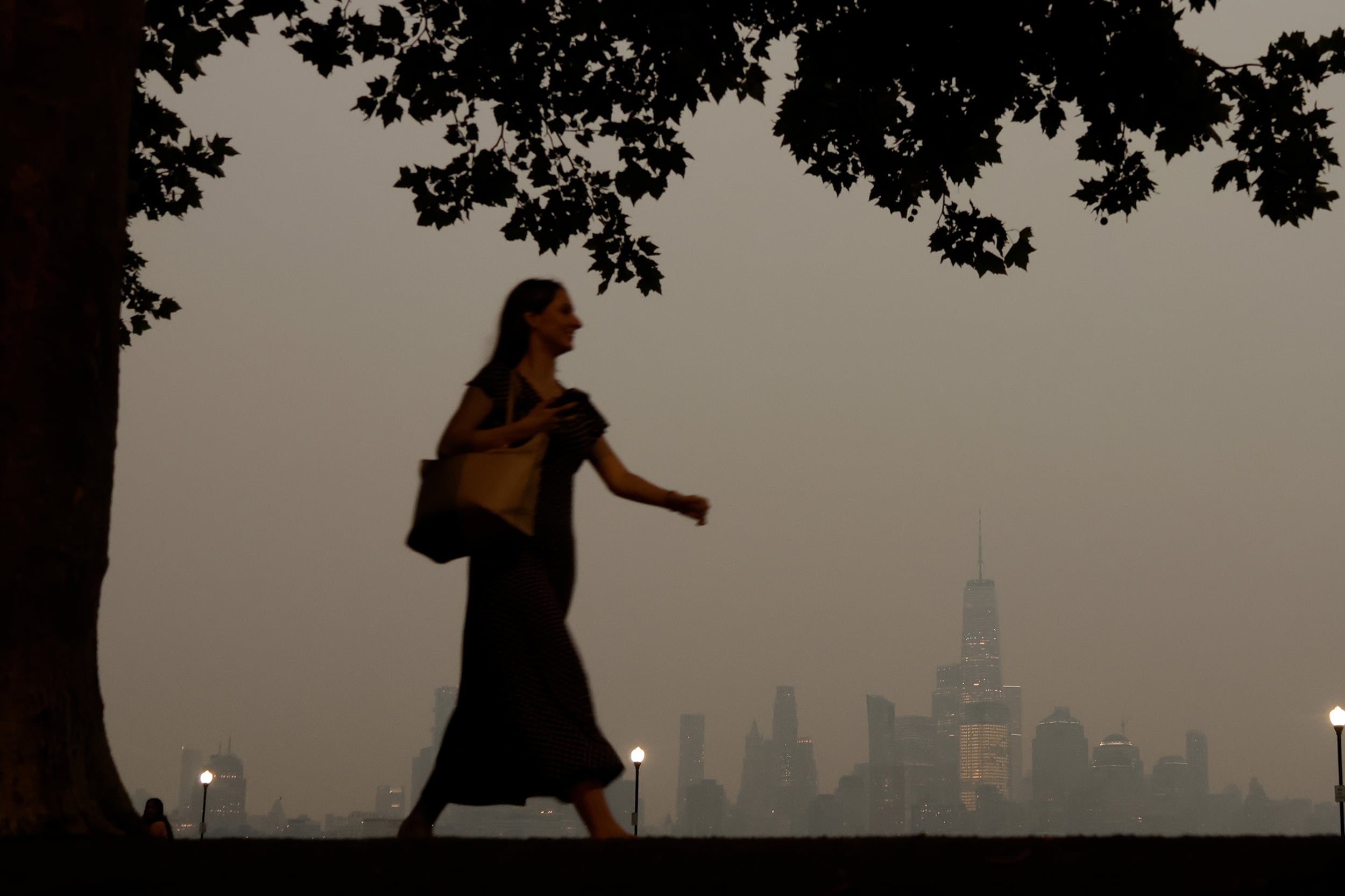 A woman walks by a tree during a wildfire, smoke-filled evening with the Manhattan skyline in the background.