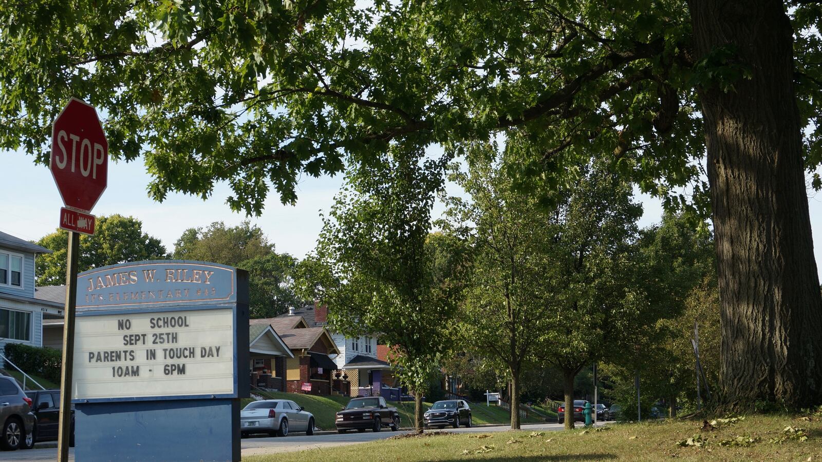 A blue school sign reading “James W. Riley” sits on a grassy lawn with a red stop sign in the foreground.