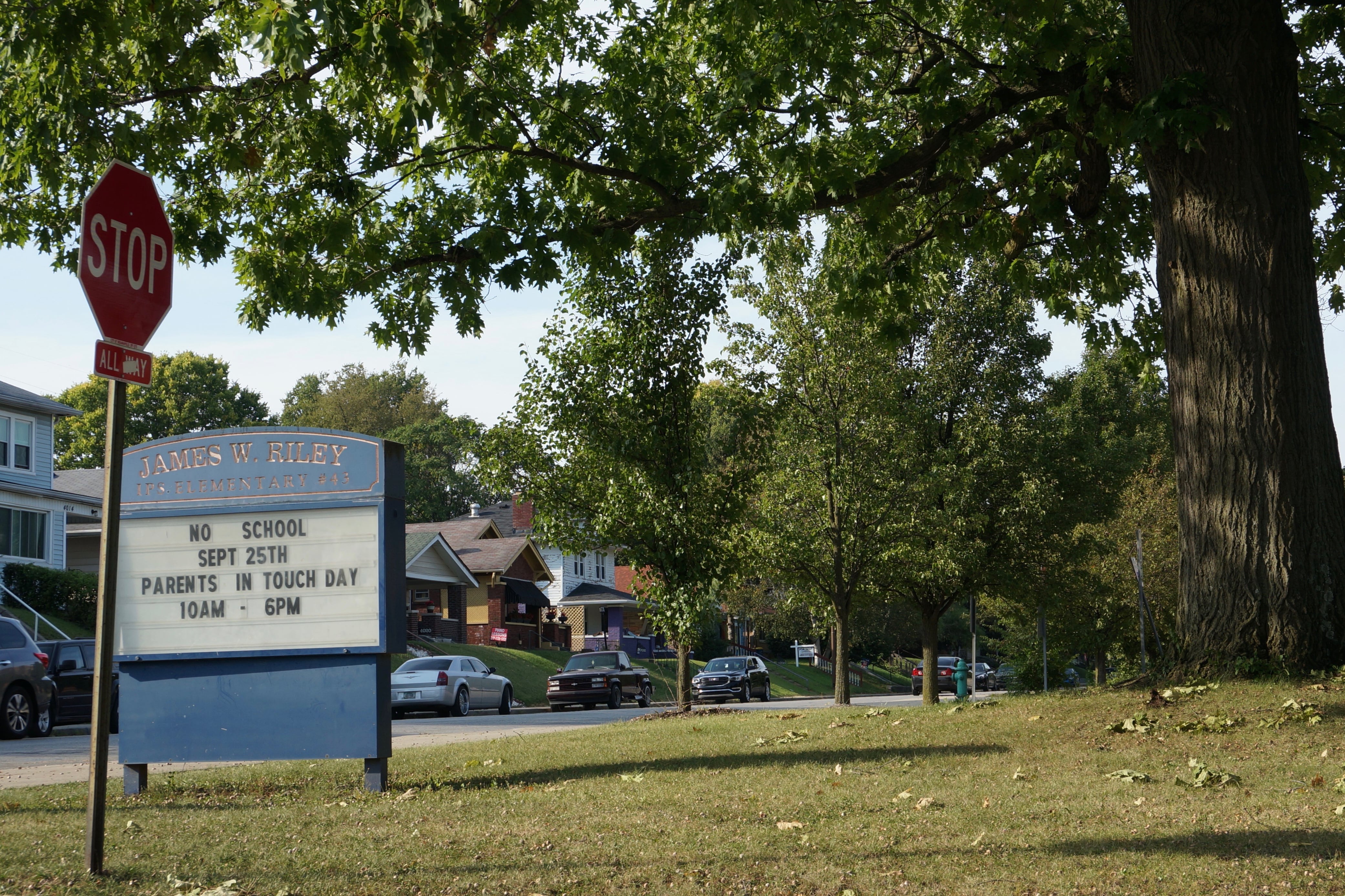 A blue school sign reading “James W. Riley” sits on a grassy lawn with a red stop sign in the foreground.