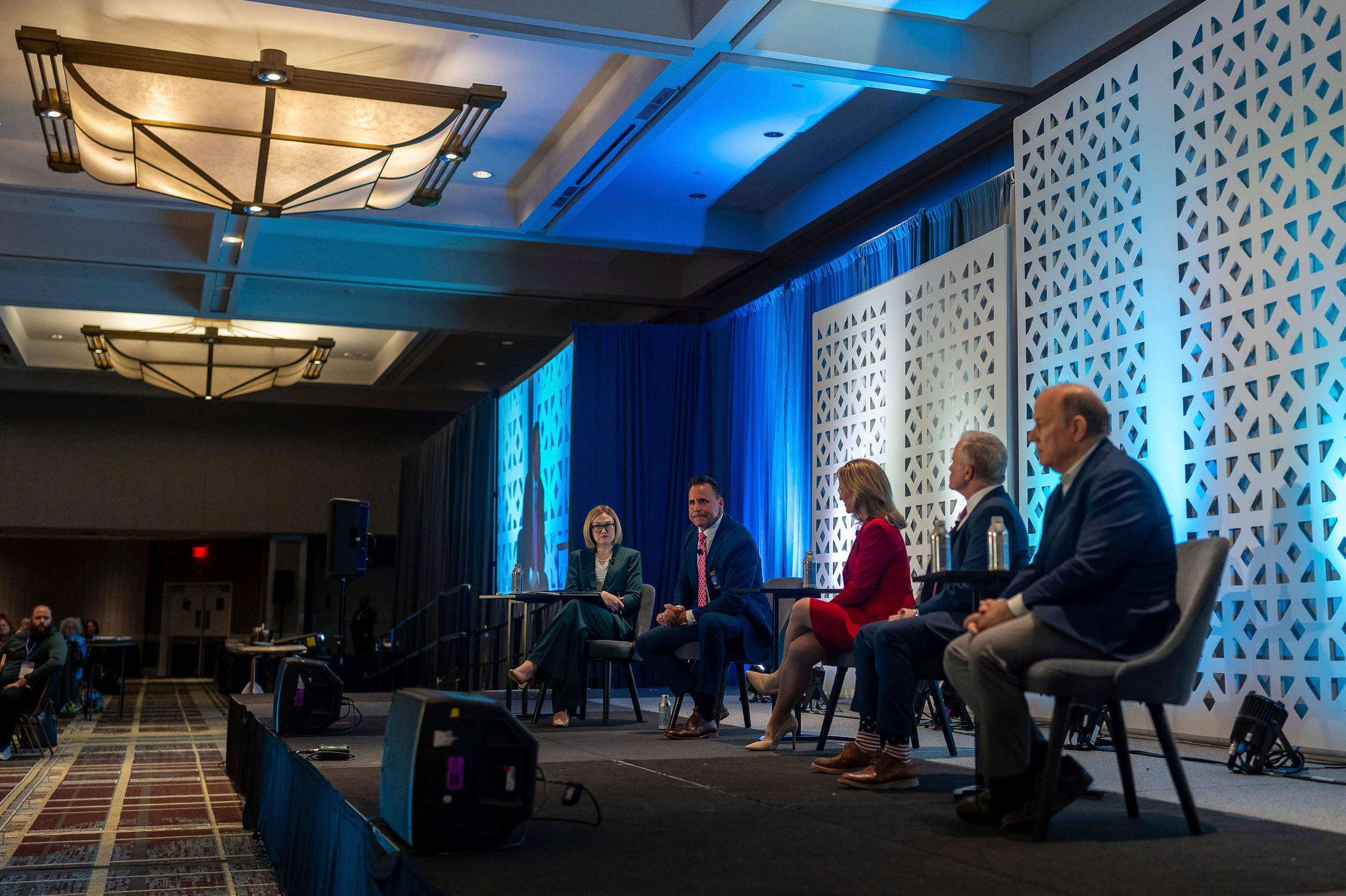 A photograph of five adults in suits sitting in chairs on a stage in a large conference room.