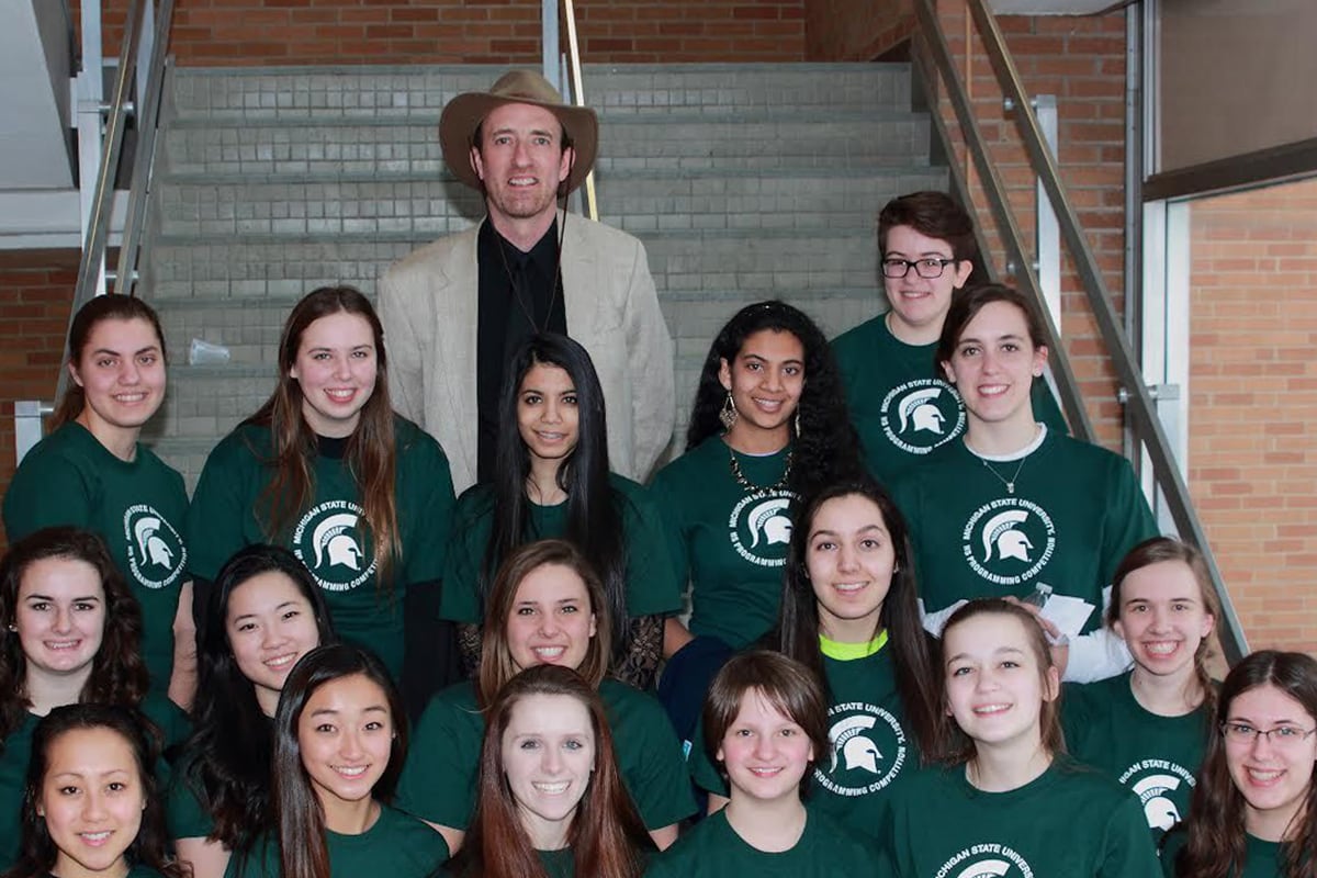 A man in a suit poses with a high school girl's programming team on a set of stairs.