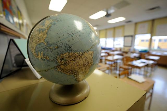 A world globe sits on the side of a table with an empty classroom full of desks in the background.