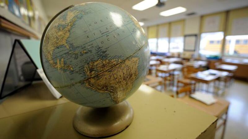 A world globe sits on the side of a table with an empty classroom full of desks in the background.