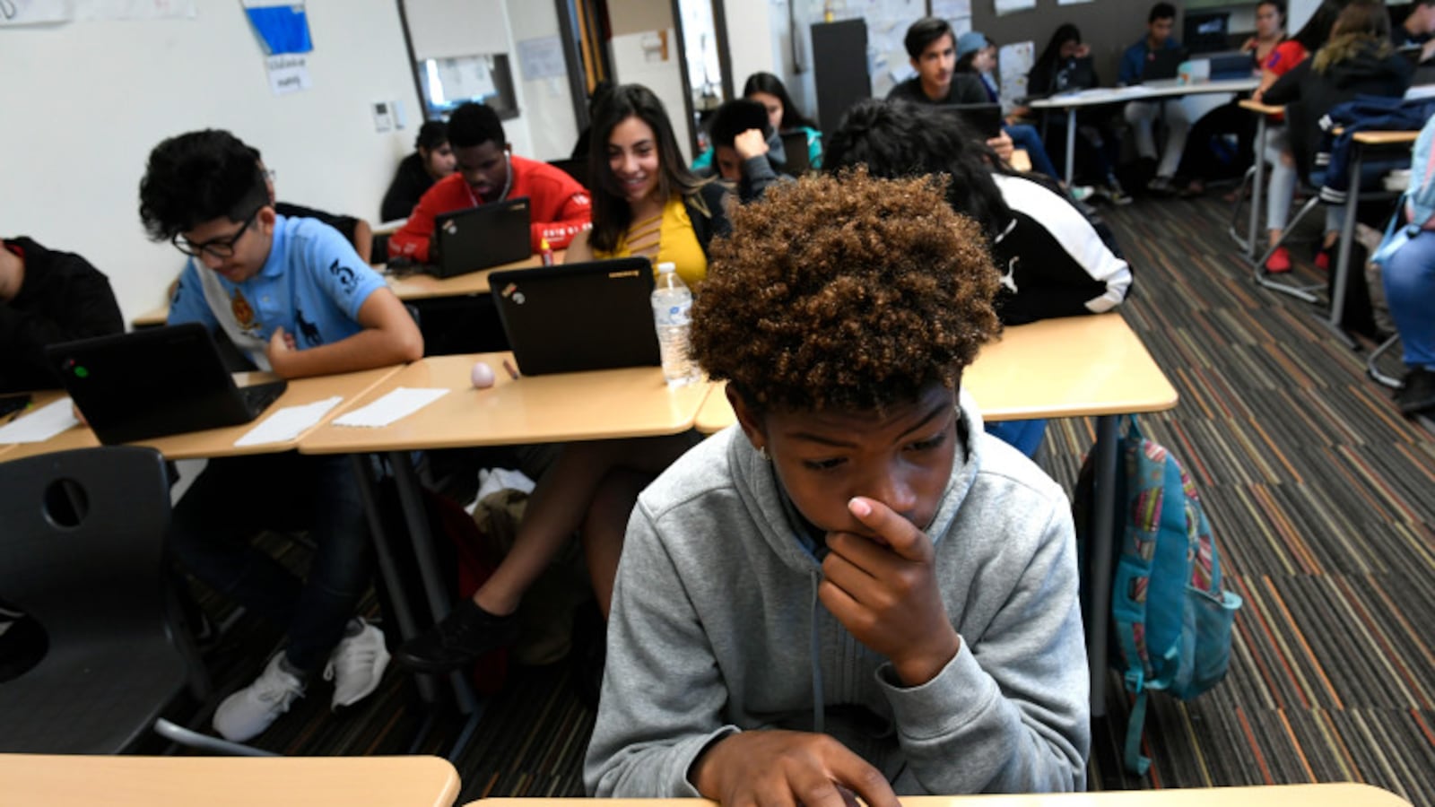 A high school student works at a computer amongst his classmates, several of which are behind him.