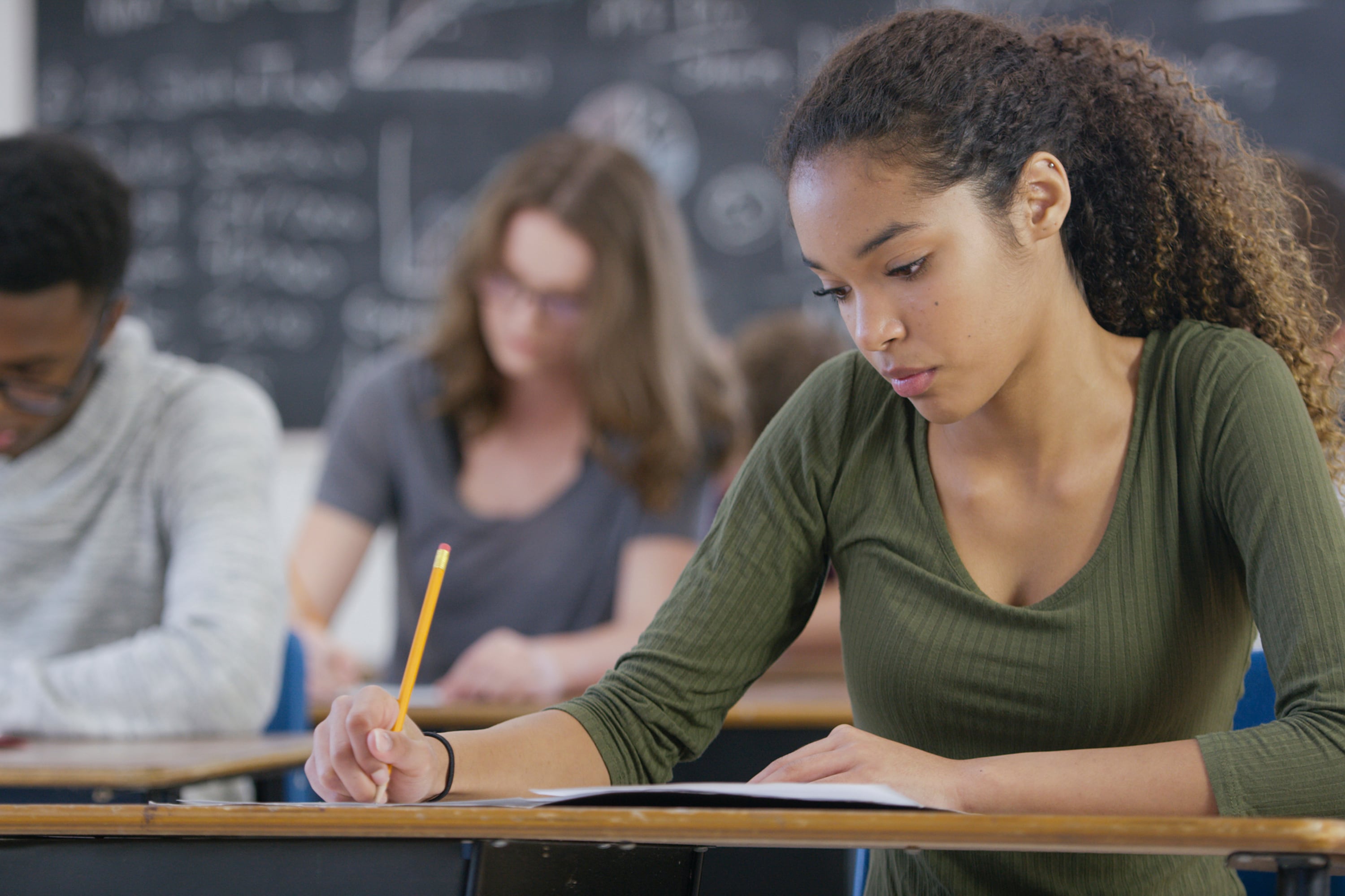A high school girl wearing a green shirt sits at a desk taking a test with two students in the background.