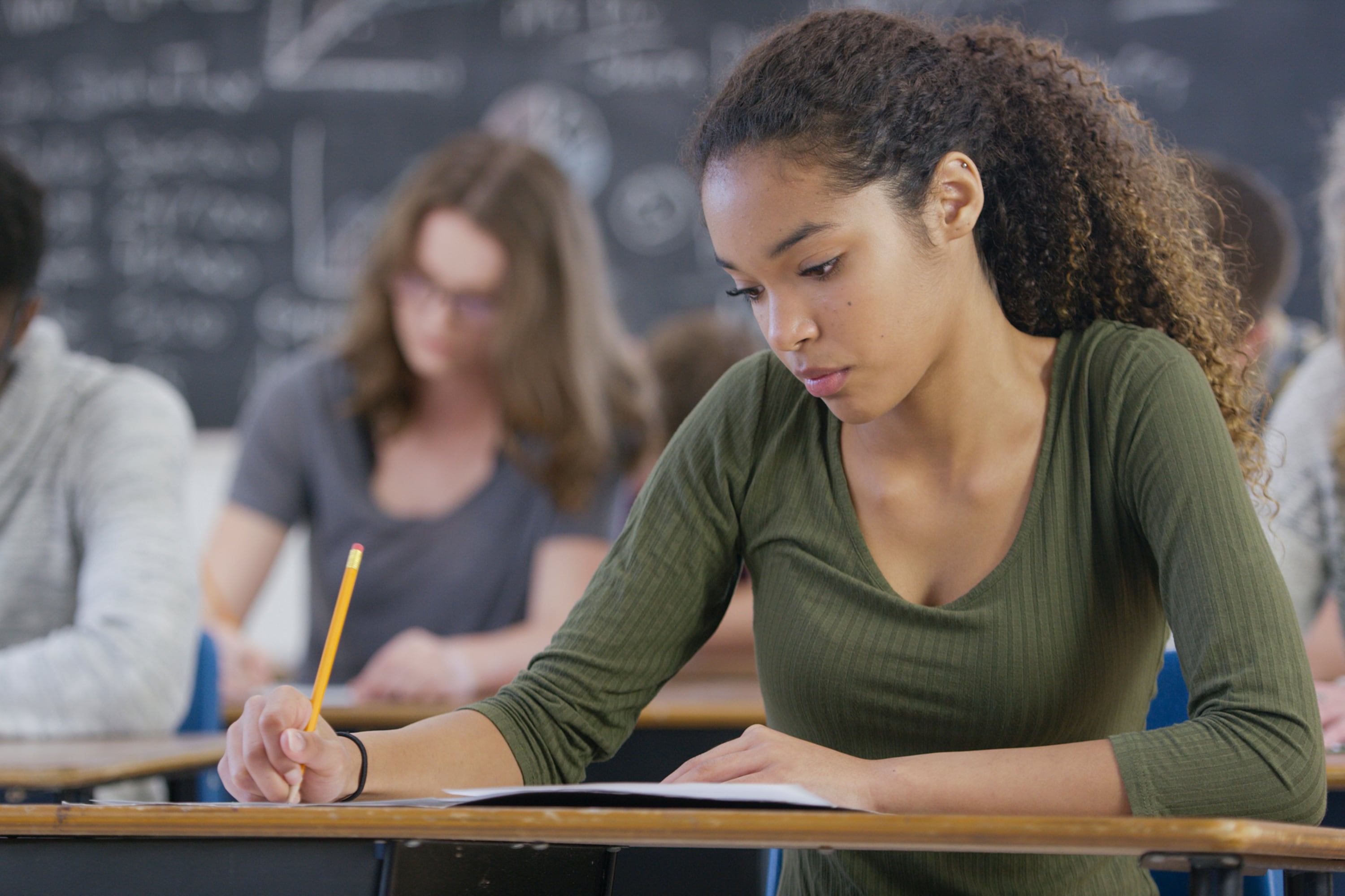 A high school girl wearing a green shirt sits at a desk taking a test with two students in the background.