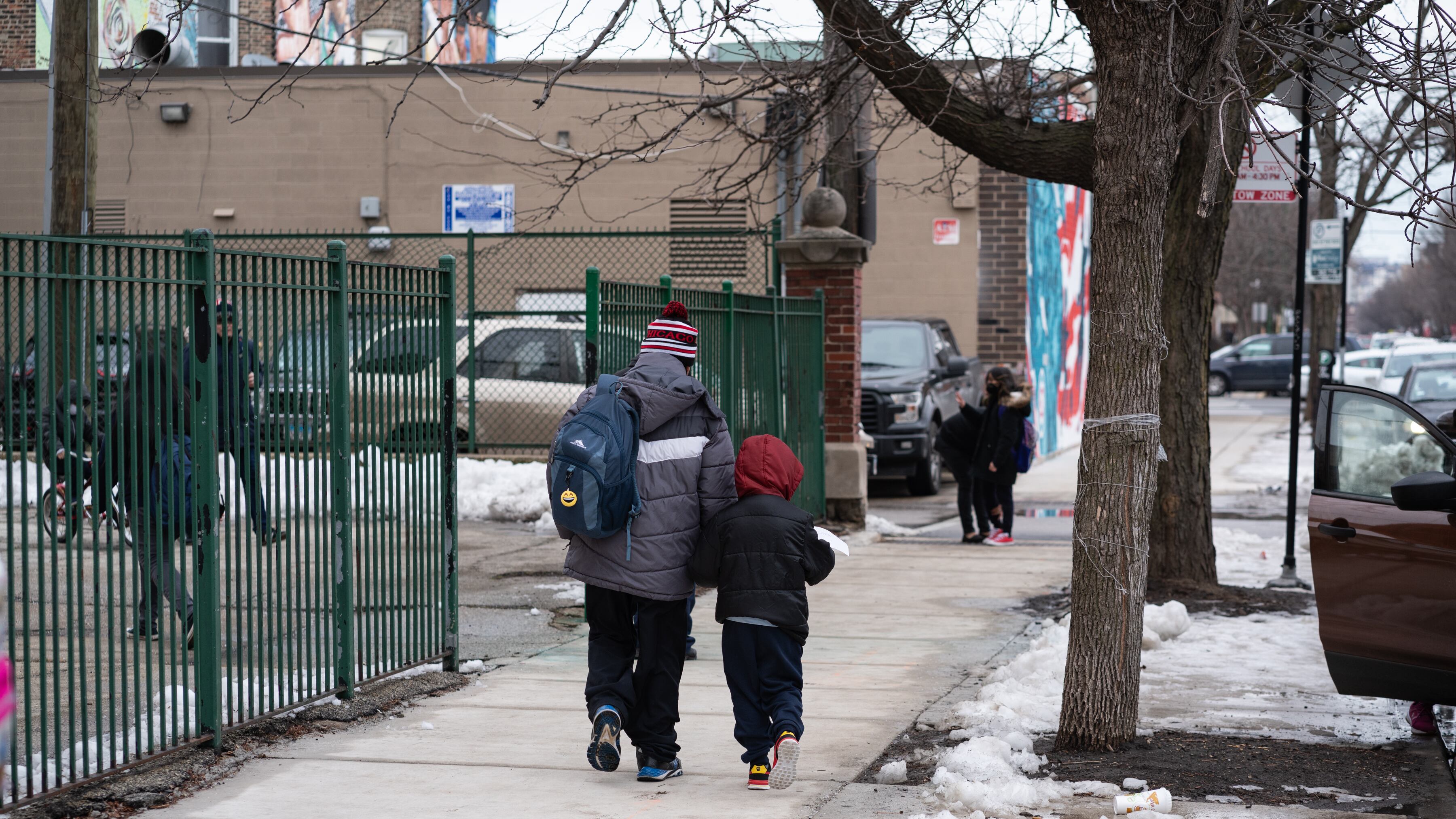 A man and a child in winter coats walk on the sidewalk by a school building.