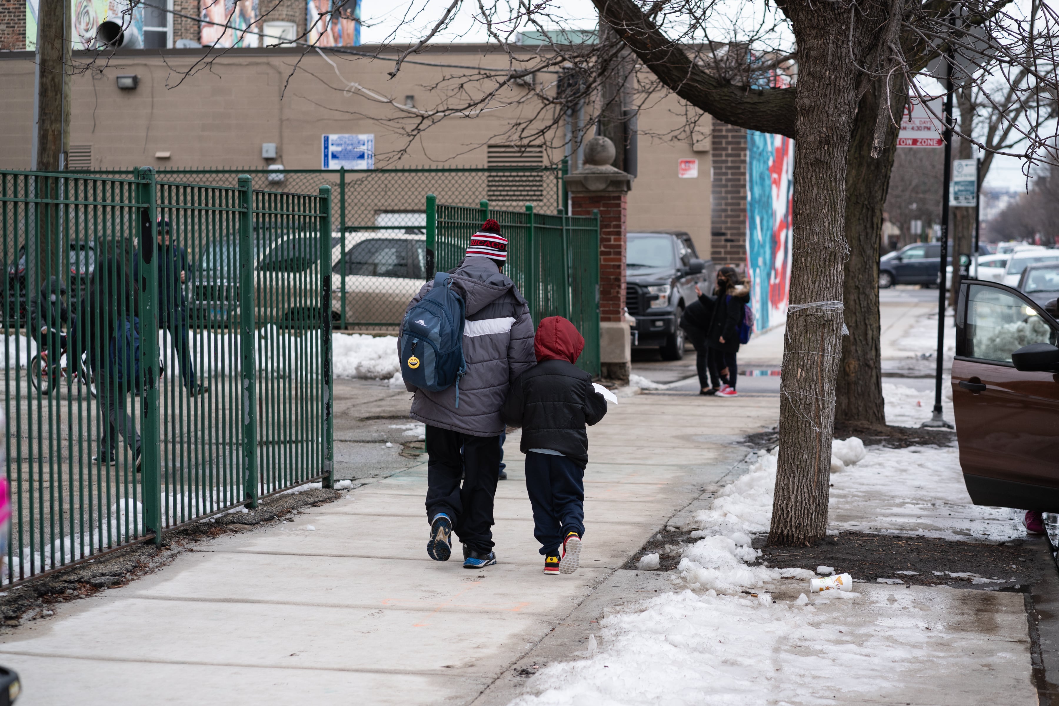 A man and a child in winter coats walk on the sidewalk by a school building.