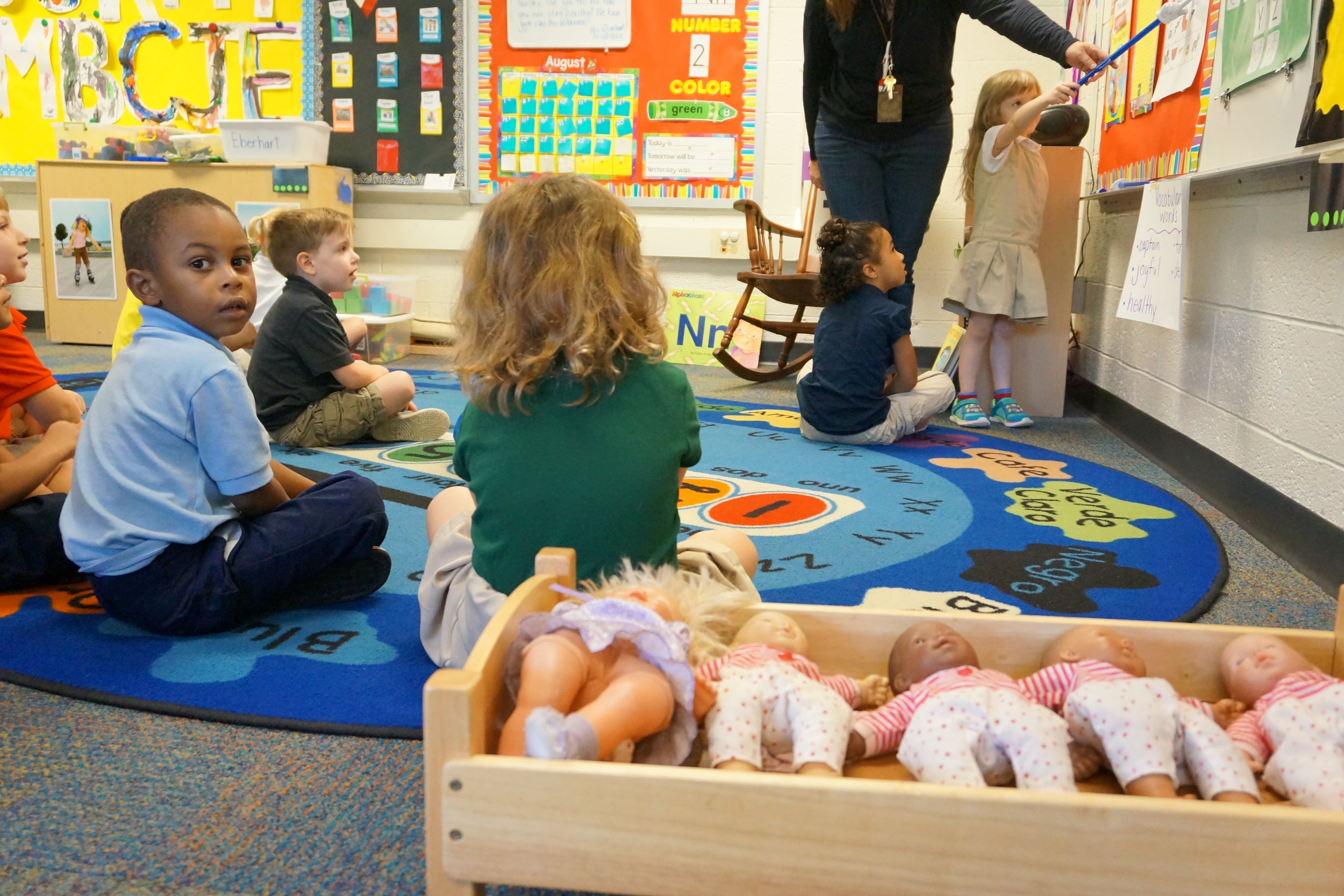A group of six preschoolers participate in an exercise while a boy in a blue shirt looks back at the photographer. There is a row of dolls sitting in a wooden bin in the foreground.