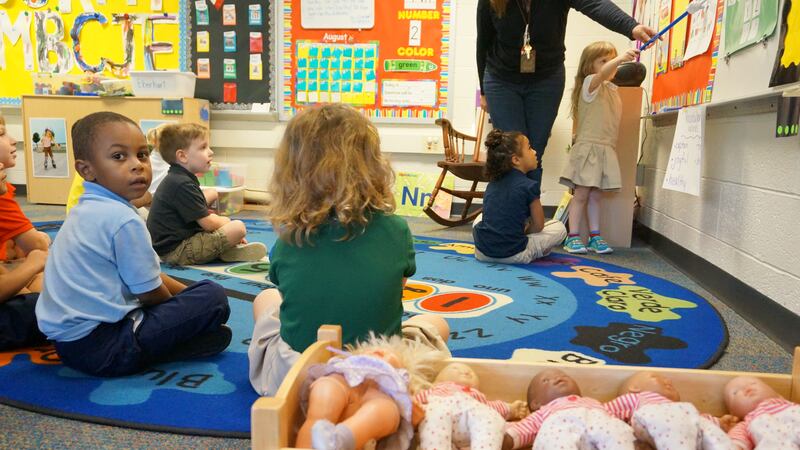 A group of six preschoolers participate in an exercise while a boy in a blue shirt looks back at the photographer. There is a row of dolls sitting in a wooden bin in the foreground.