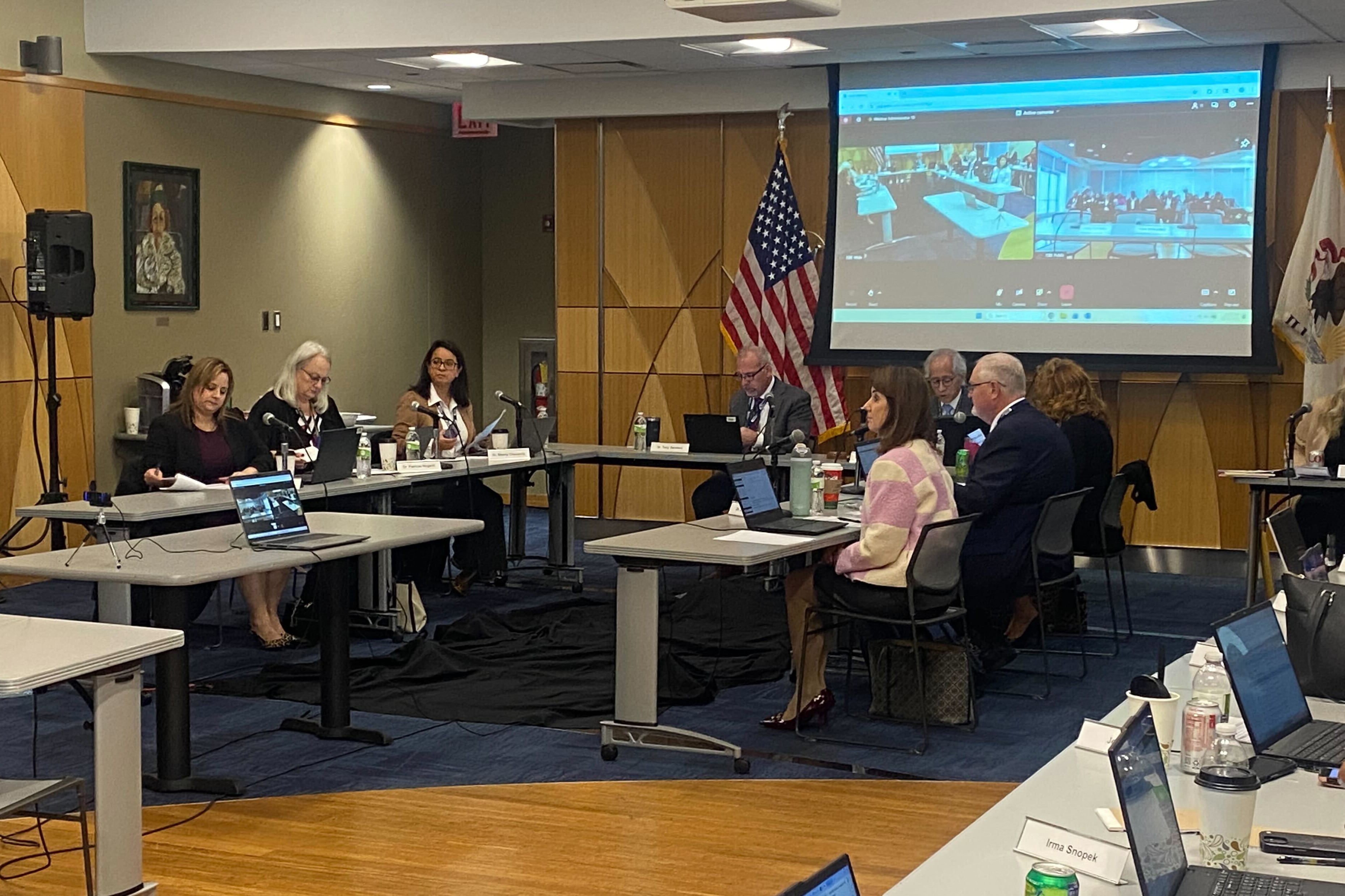 A photograph of a group of adults in business clothes sit around tables in a meeting room.