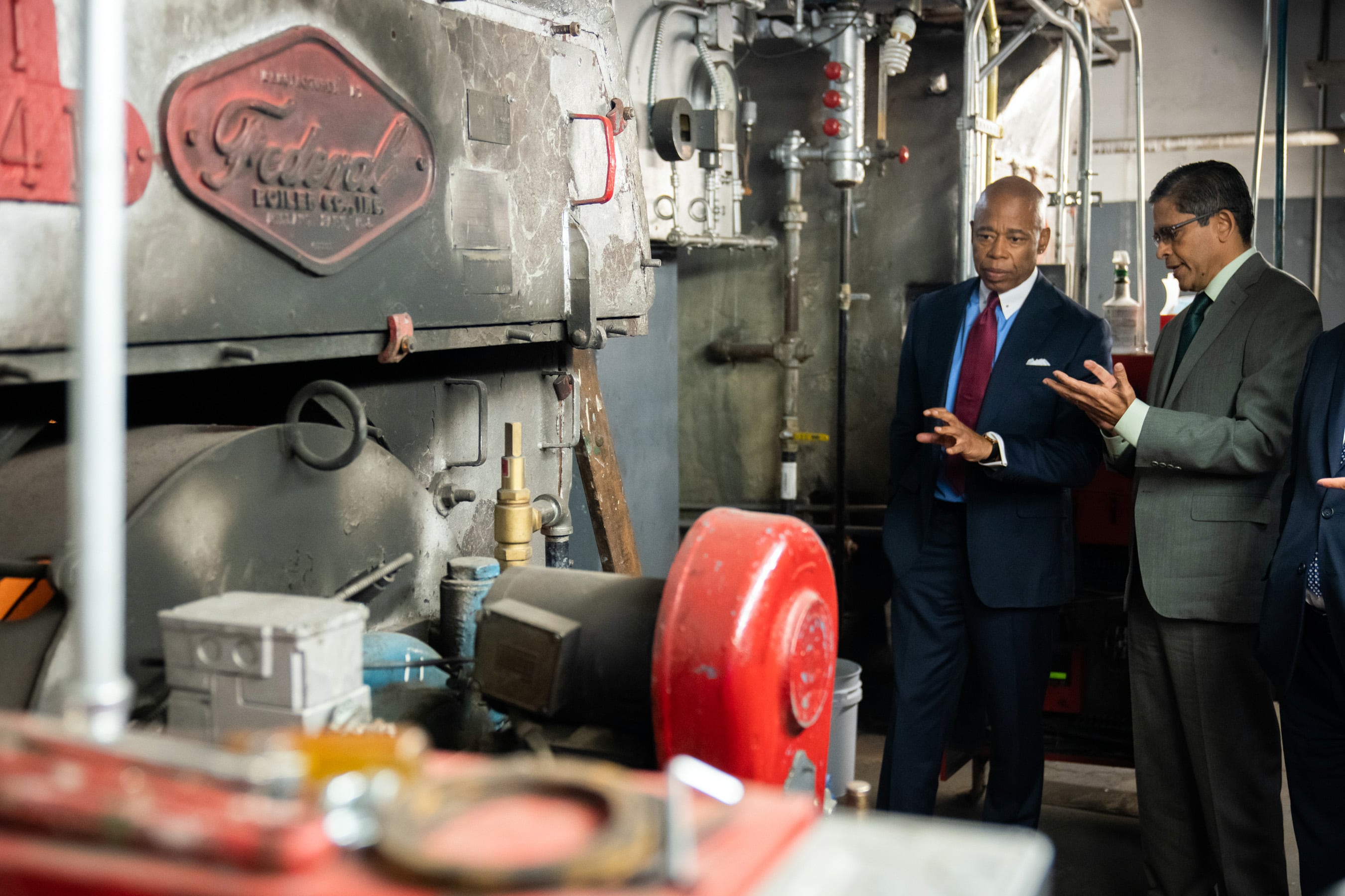 Two men stand in front of an old boiler in a school basement.