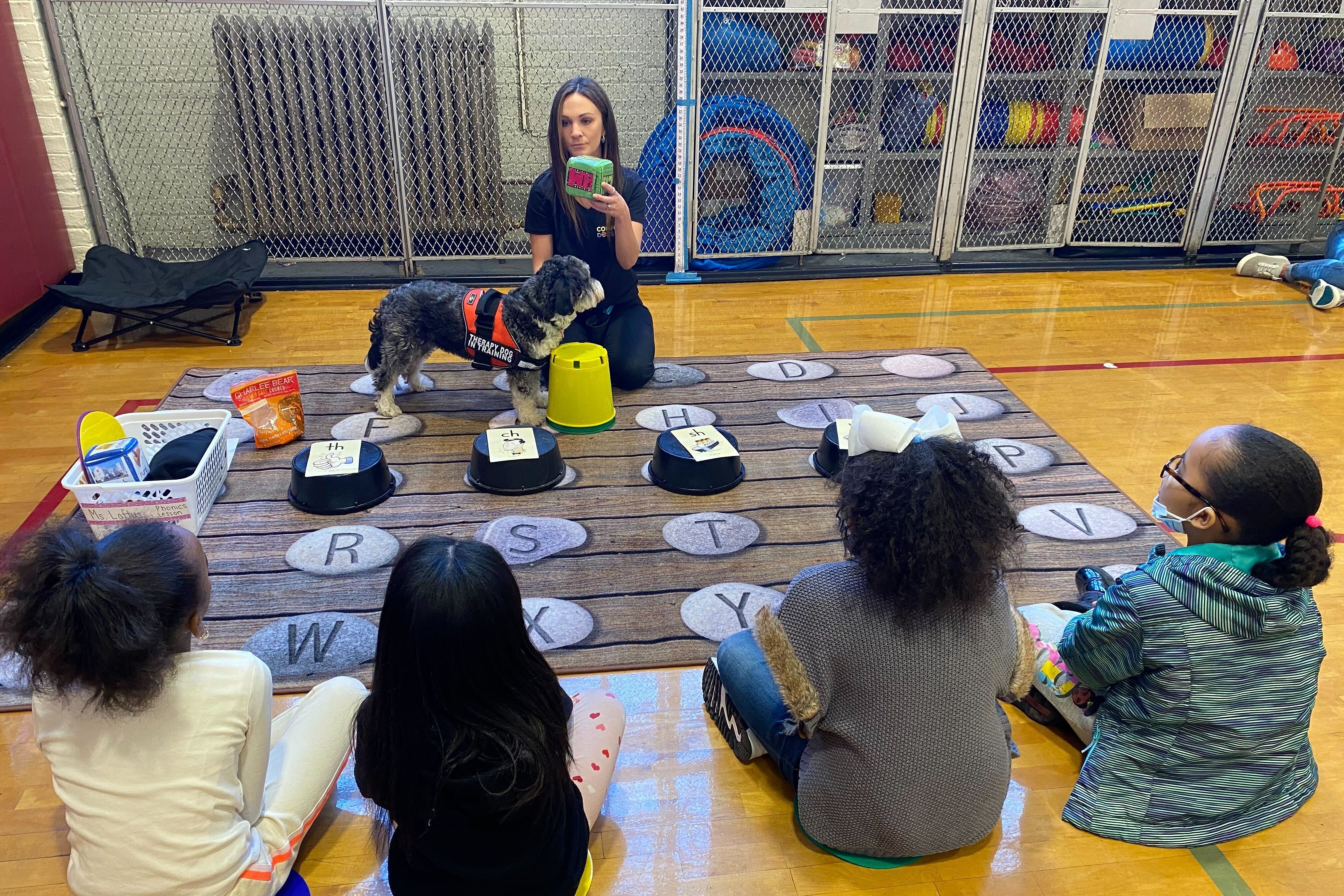 A woman holds a die as a dog stands in front of her and four children sit across from her.