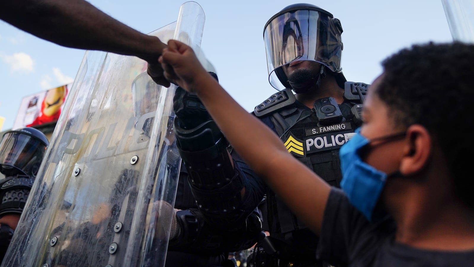 A young boy in a blue protective mask raises his fist as a protester bumps his fist with a police officer in full riot gear, surrounded by several other police.